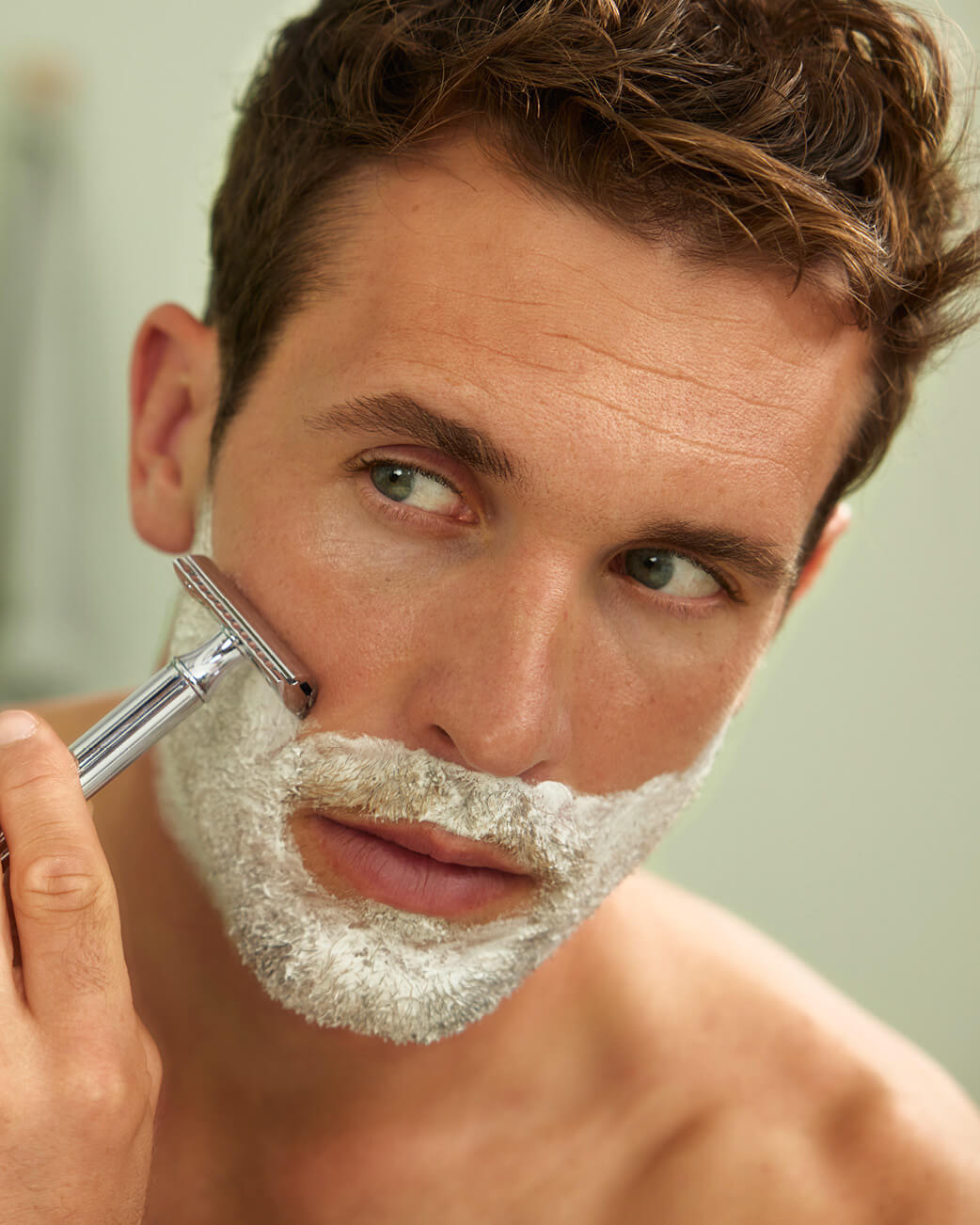 Man with brown hair using a safety razor to shave his face.