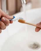 Person applying toothpaste to a bamboo toothbrush over a blurred bathroom sink background.