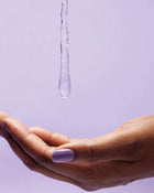 Hand with purple nail polish holding a droplet of water against a light purple background.