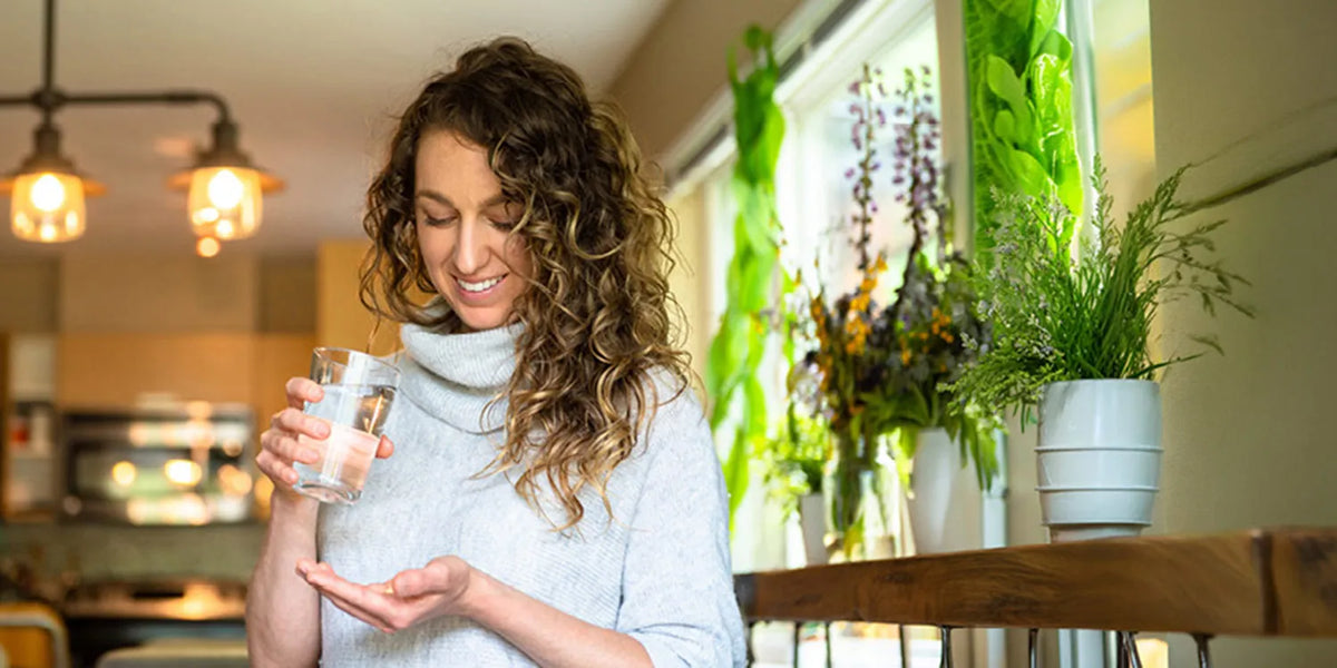A smiling woman with curly brown hair stands in a bright kitchen or cafe setting, holding a glass of water and looking down at it. She is wearing a light gray turtleneck sweater, with potted plants and warm hanging lights in the background.