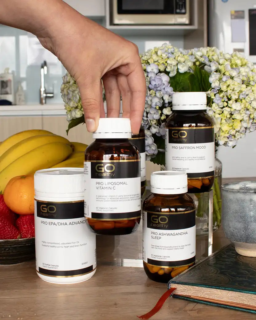 Hand holding a bottle of GO Healthy supplements on a kitchen counter with various bottles and fruits in the background.