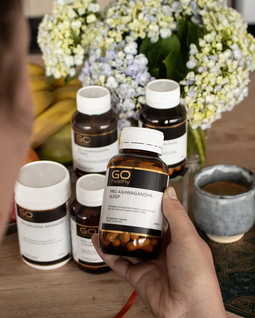 Hand holding a bottle of GO Healthy Pro Ashwagandha Sleep supplement with other supplements and flowers in the background.