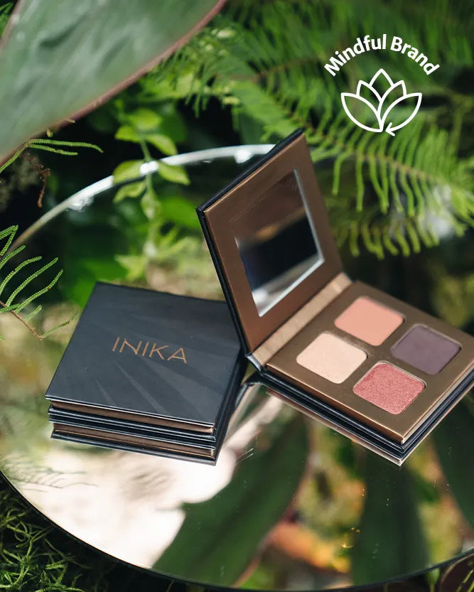 An open INIKA eyeshadow palette with four shades and a mirror, resting on a reflective surface next to its closed black case, with green foliage in the background and a 'Mindful Brand' logo overlay.