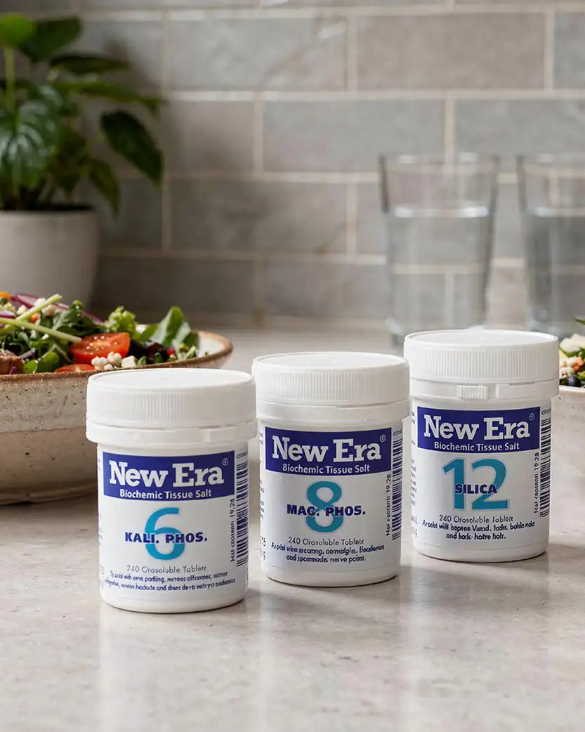 Three containers of New Era Biochemical Tissue Salt on a kitchen counter with a salad and glasses in the background.