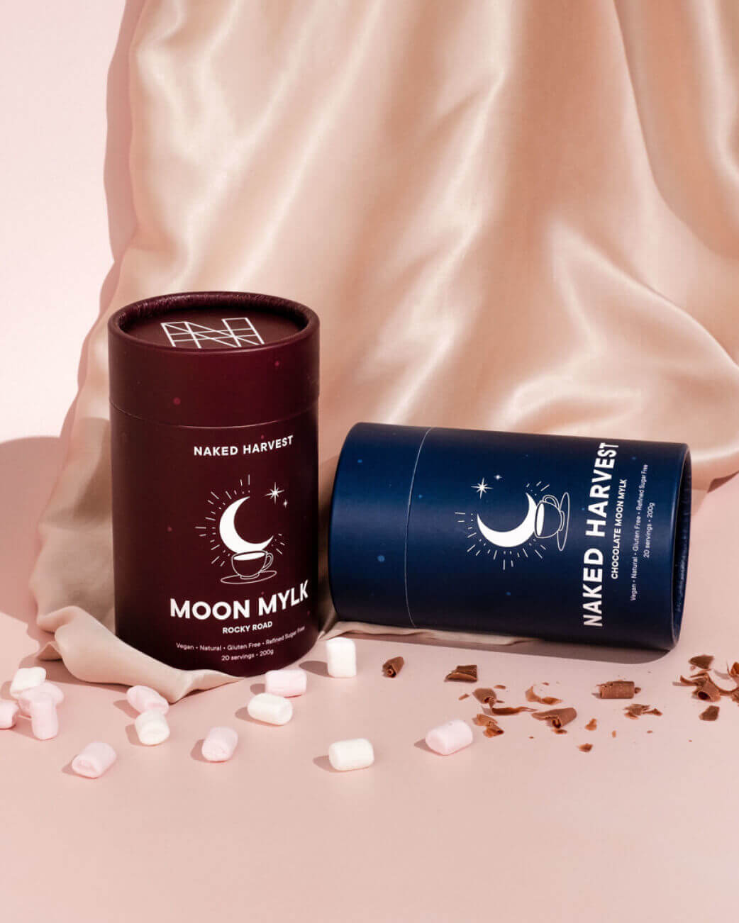 Two cylindrical Naked Harvest product containers on a soft pink background with scattered candies.