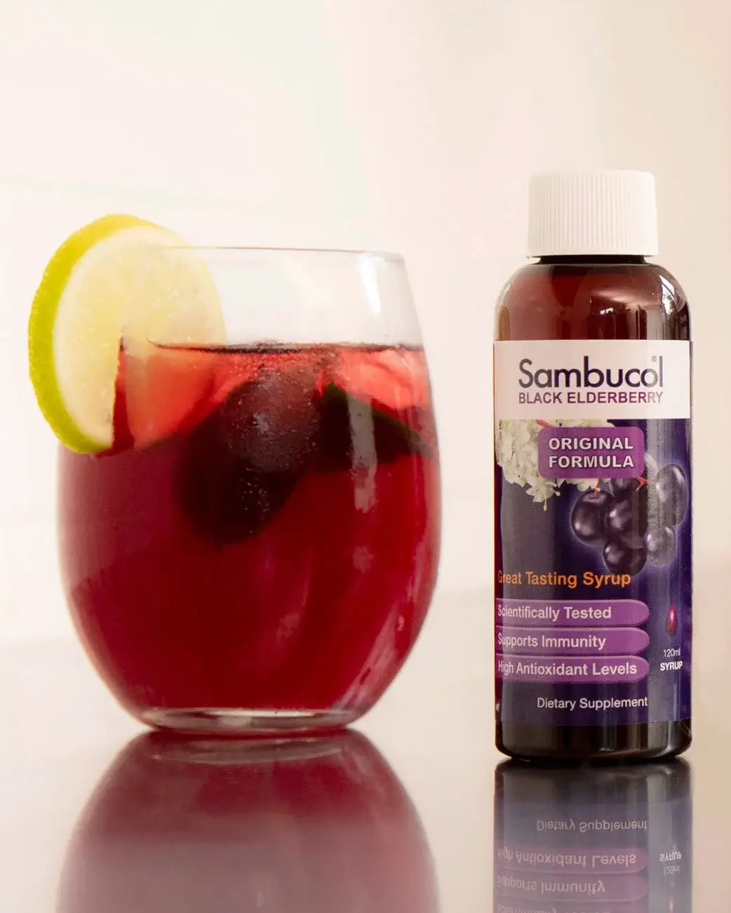 Glass of red liquid with a lemon slice next to a bottle of Sambucol Black Elderberry syrup.