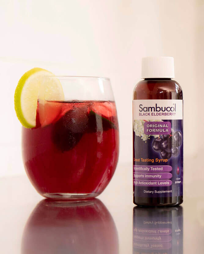 Glass of red liquid with a slice of lemon and a bottle of Sambucol Black Elderberry syrup on a reflective surface.