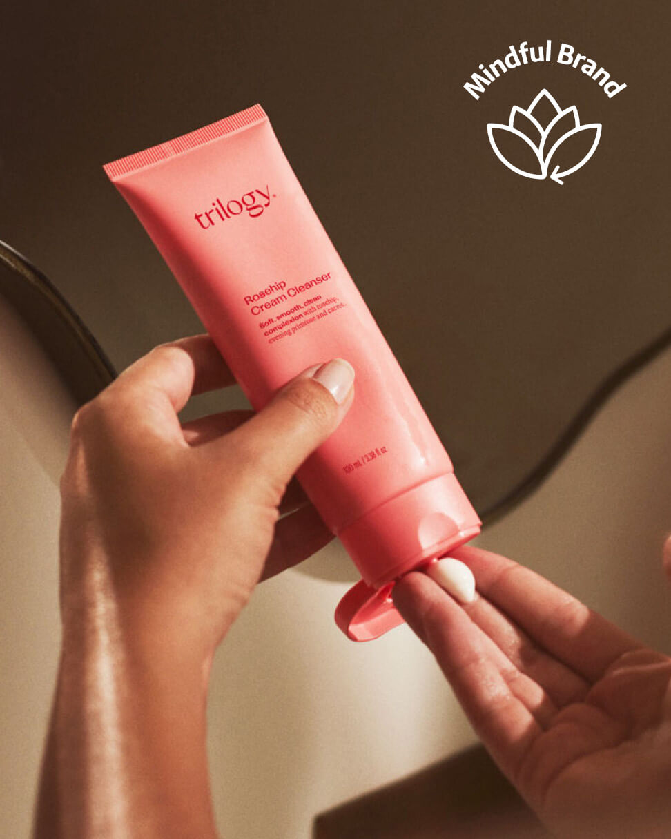 Hand holding a pink tube of Trilogy product with 'Mindful Brand' logo.