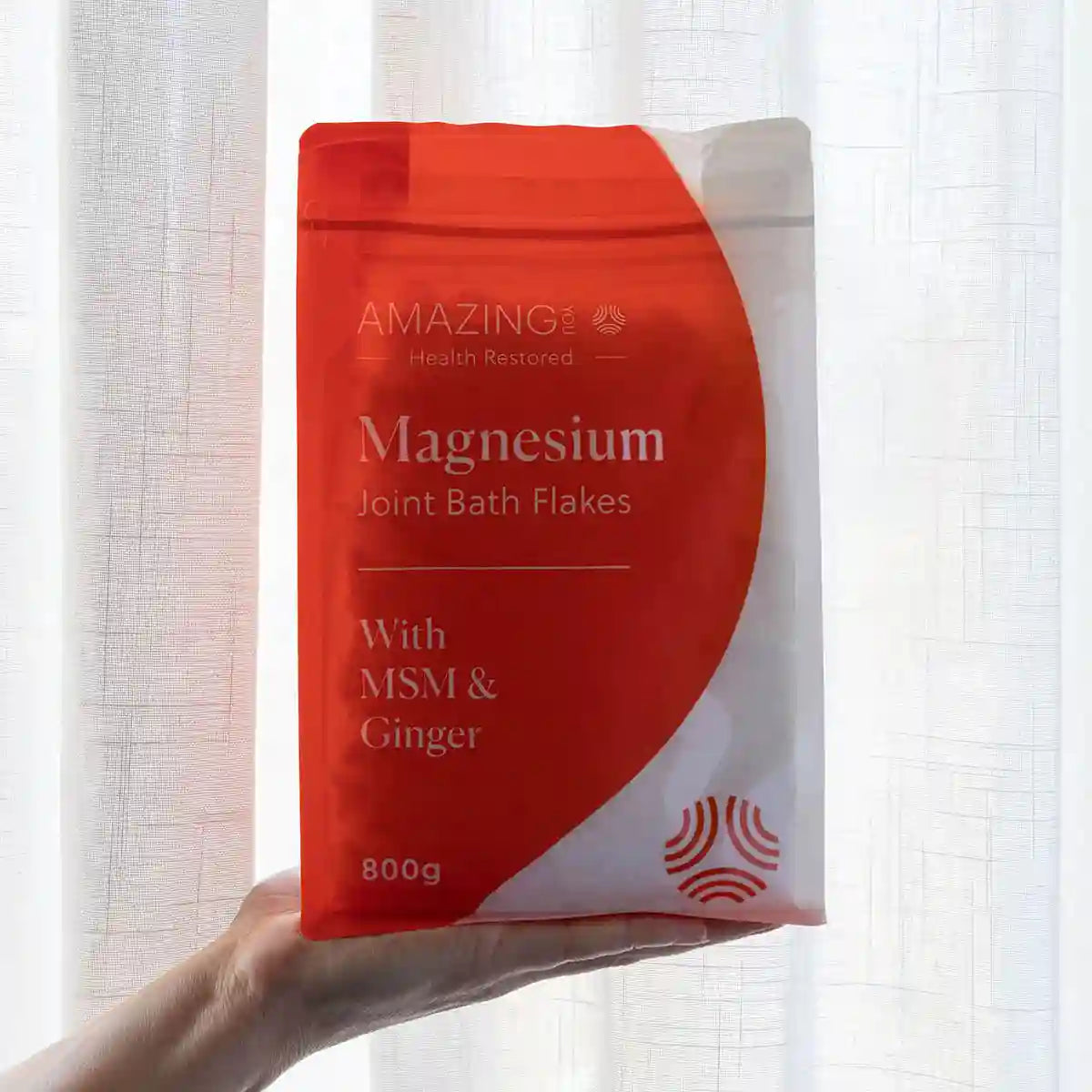 Hand holding a package of Magnesium Joint Bath Flakes with MSM & Ginger against a light curtain background.