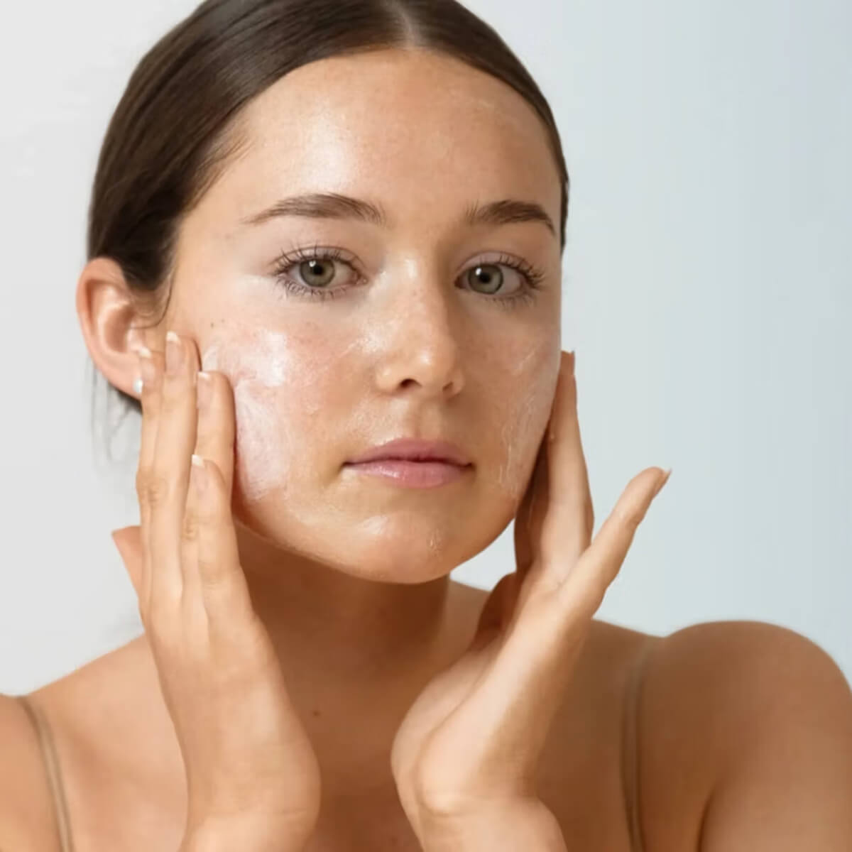 A close-up of a woman with clear, glossy skin applying product onto her cheek with both hands.