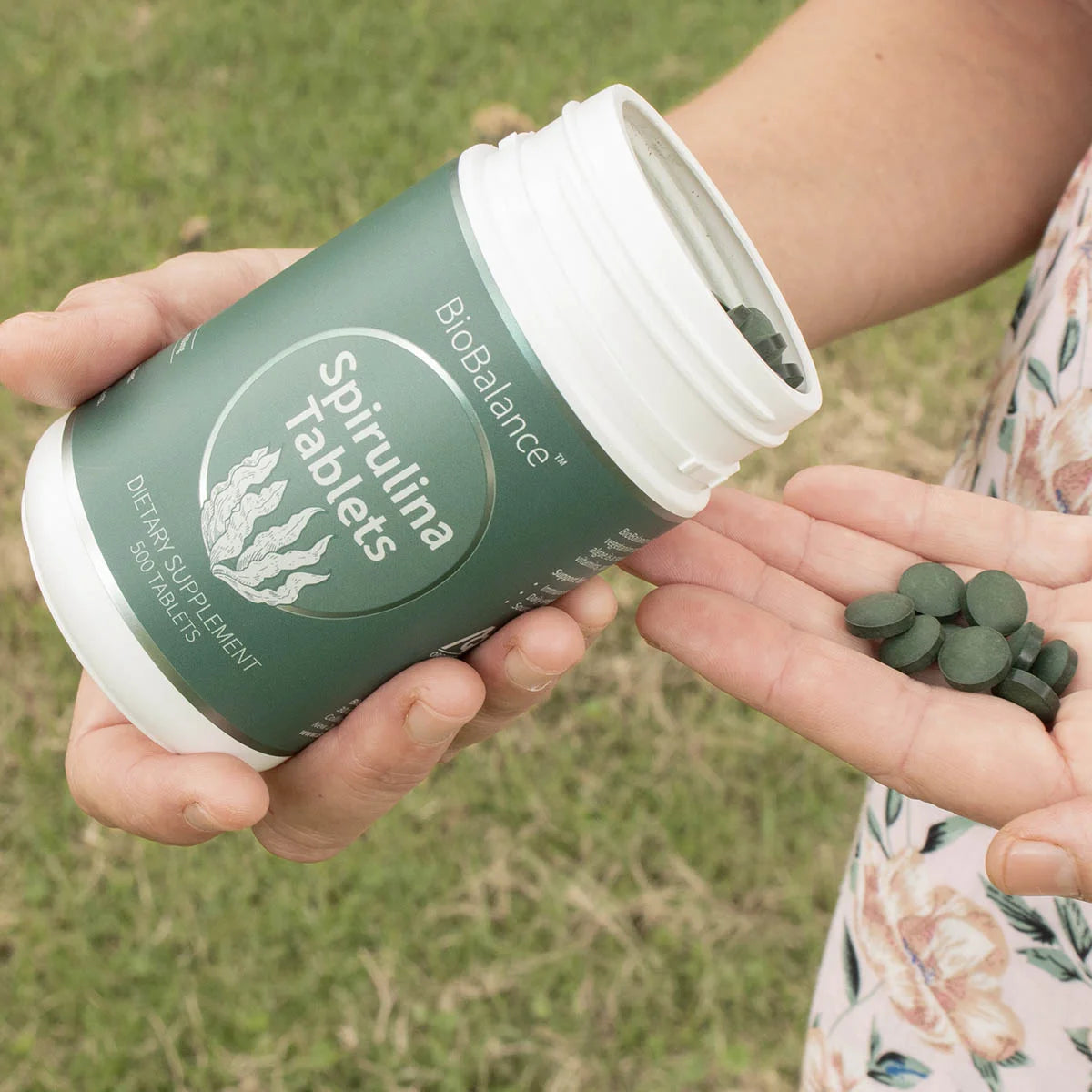 Person holding a container of BioBalance Spirulina Tablets with tablets in their hand outdoors.