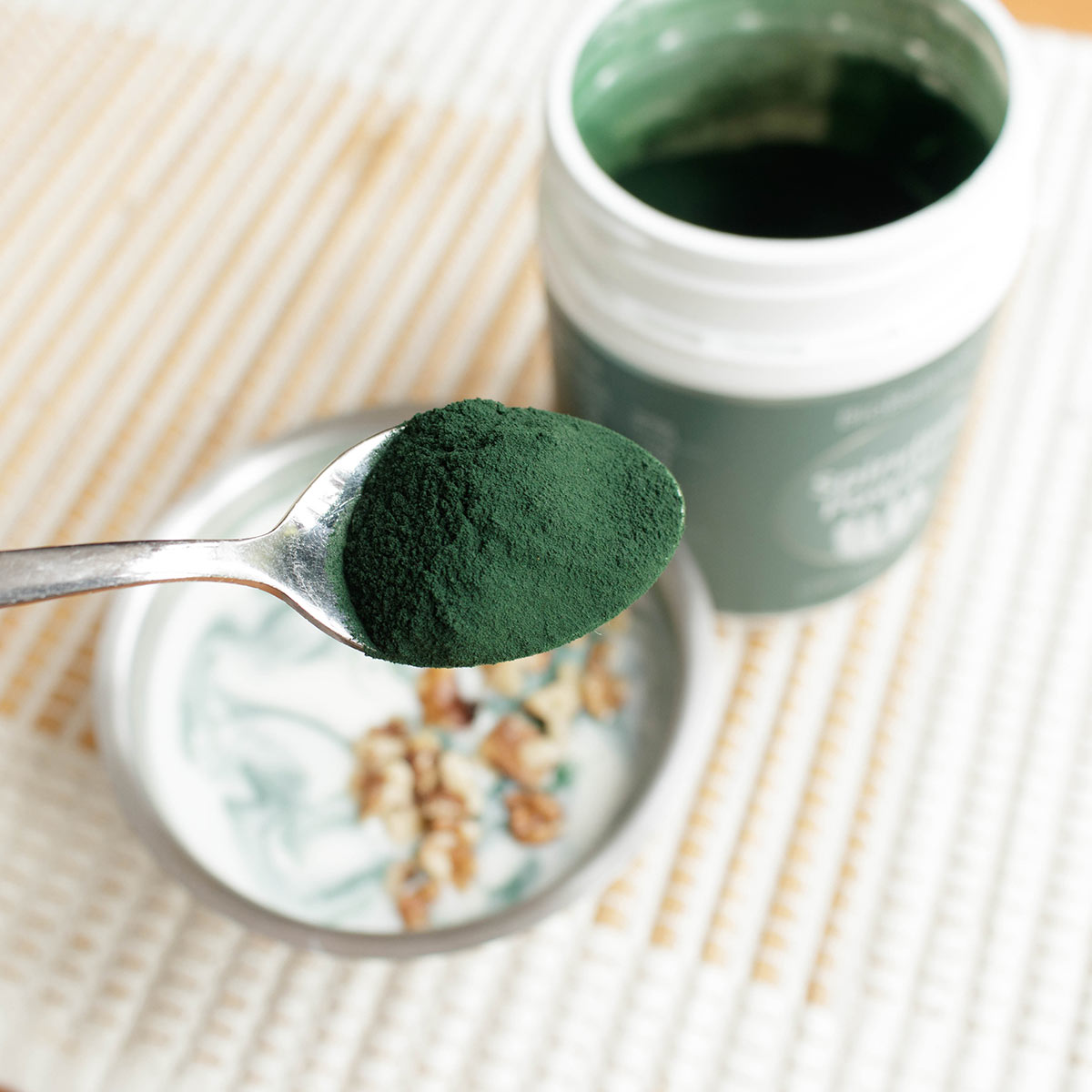 Spoonful of green spirulina powder over a bowl with open container in the background.