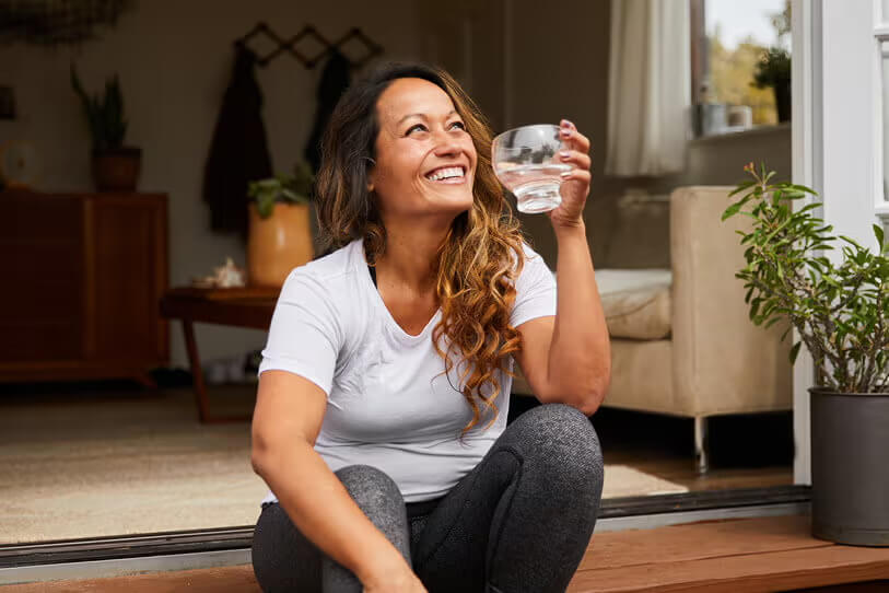 Woman sitting on a wooden step with a glass of water and smiling up at the sky.