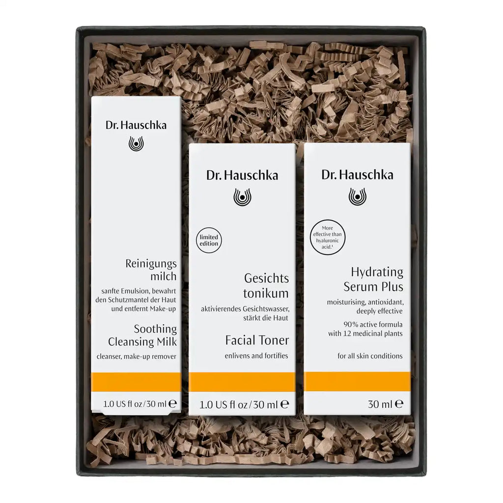 A Dr. Hauschka gift set containing three skincare bottles (Soothing Cleansing Milk, Facial Toner, and Hydrating Serum Plus) in a black box filled with brown shredded paper packaging.