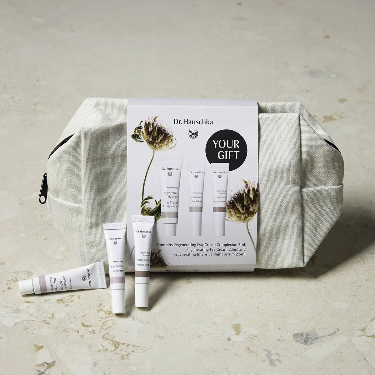 Dr. Hauschka 'Your Gift' skincare set featuring a beige canvas zippered pouch, three sample tubes, and a printed card, arranged on a textured beige stone surface.