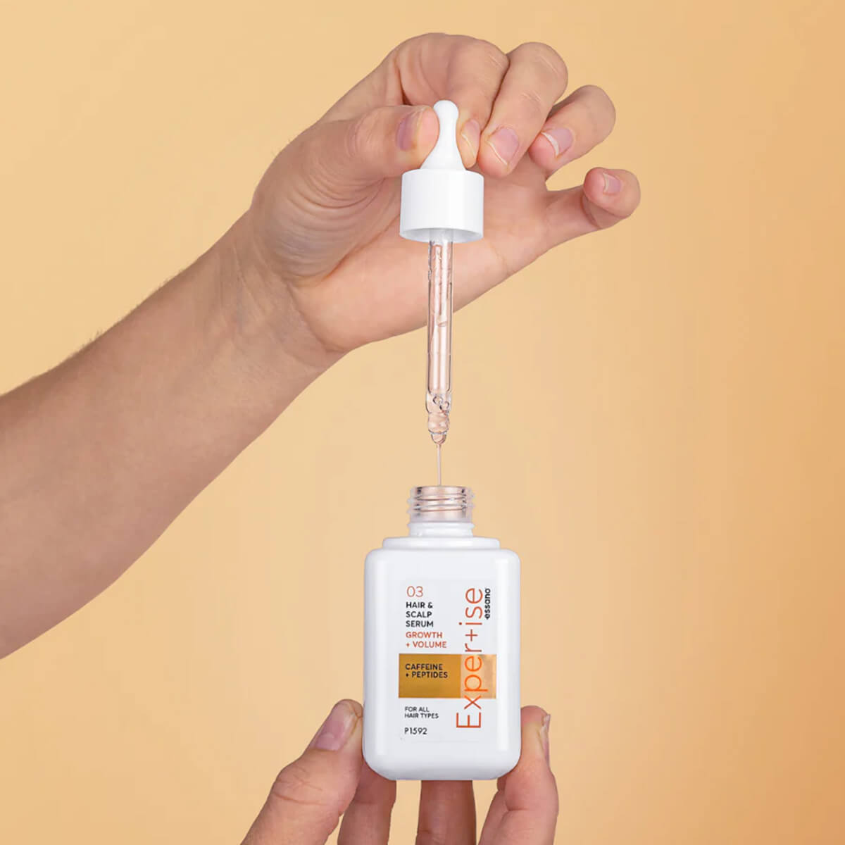 Hand holding a bottle of hair serum with a dropper against a beige background.