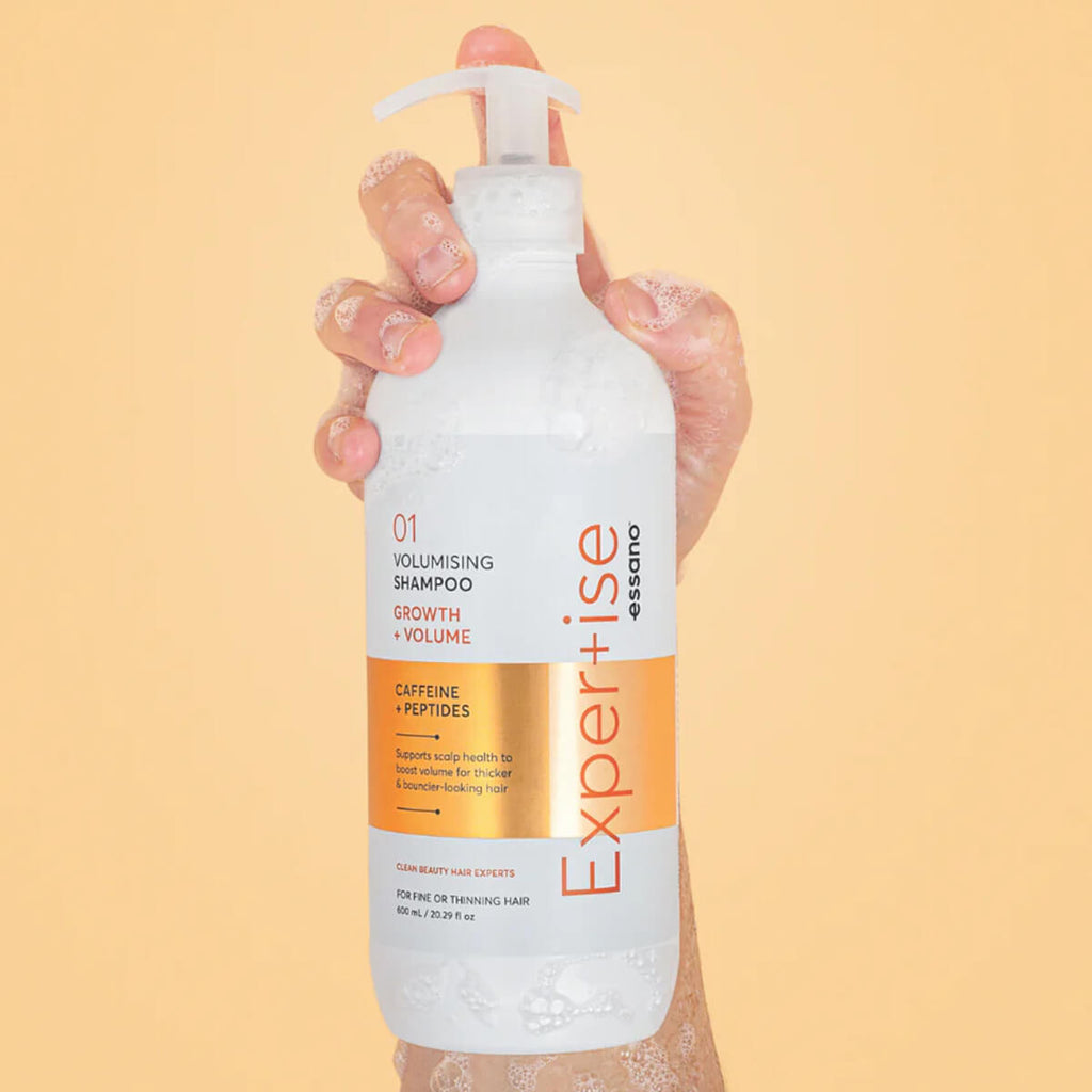 Hand holding a bottle of 'Expertise' volumising shampoo against a beige background.