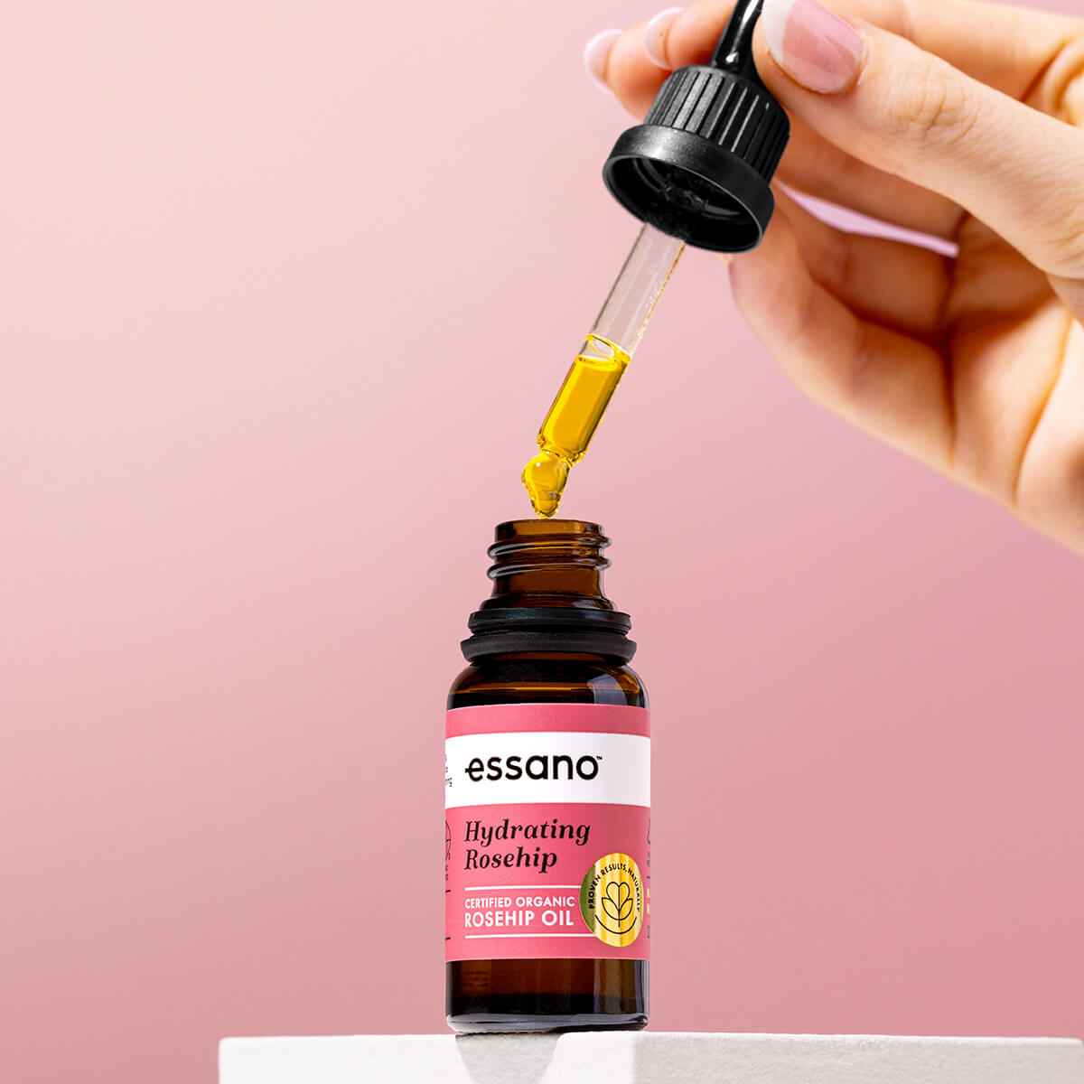 Essano dropper bottle with a hand dropping oil onto it against a pink background.
