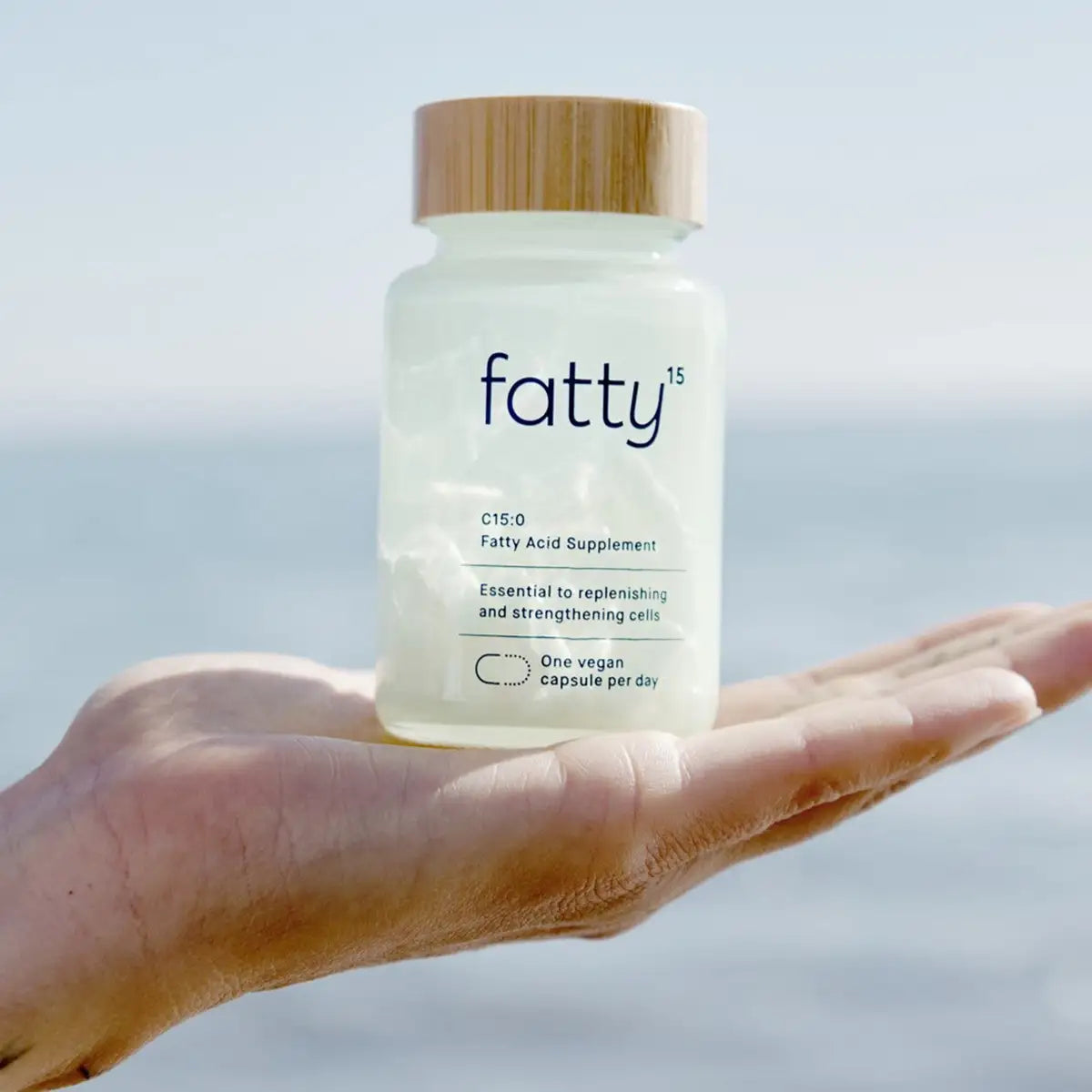 A hand holding a frosted supplement bottle labeled 'fatty15' with a bamboo-style lid, set against a soft blurred outdoor background.