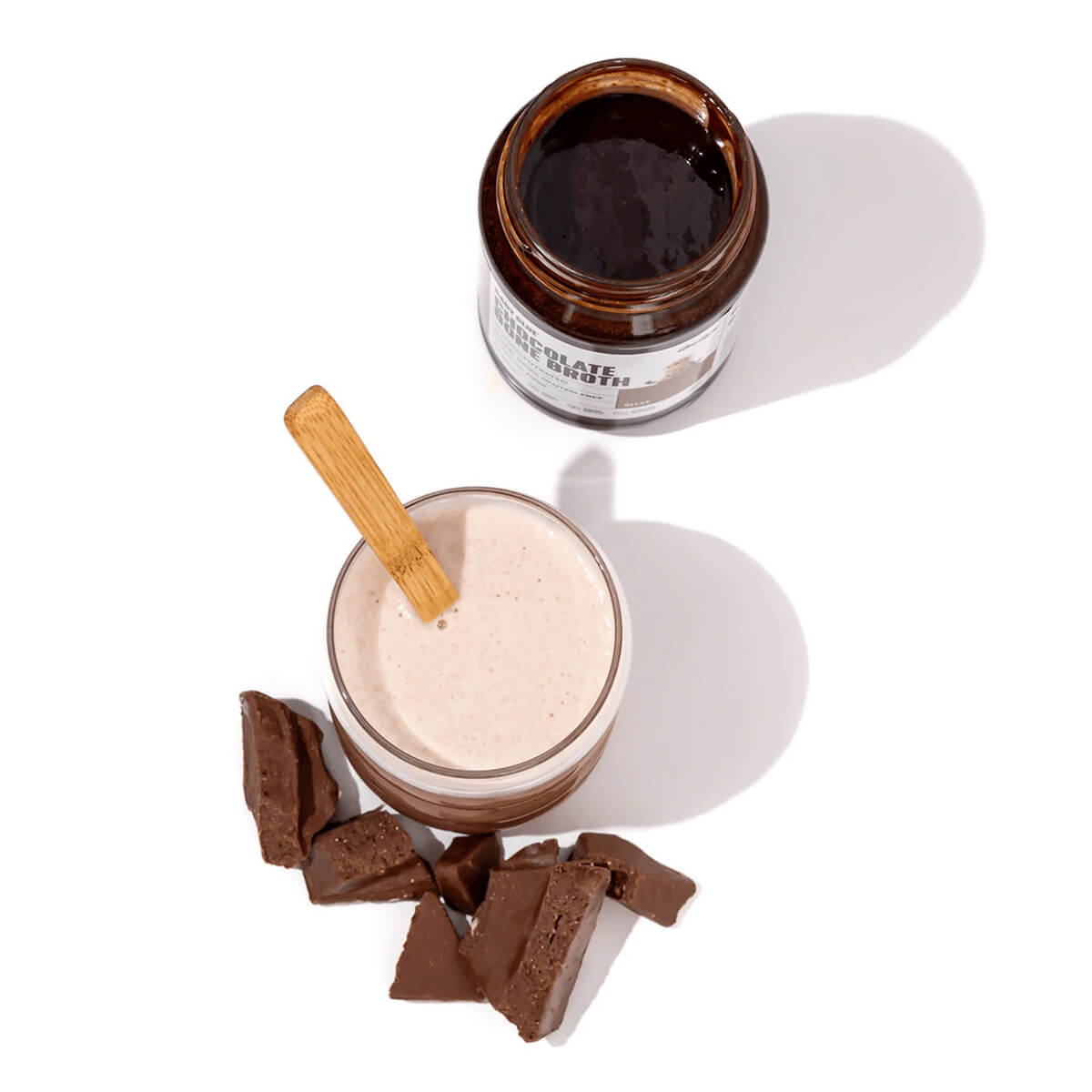 Glass of chocolate milkshake with a wooden spoon, jar of chocolate bone broth, and chocolate pieces on a white background.