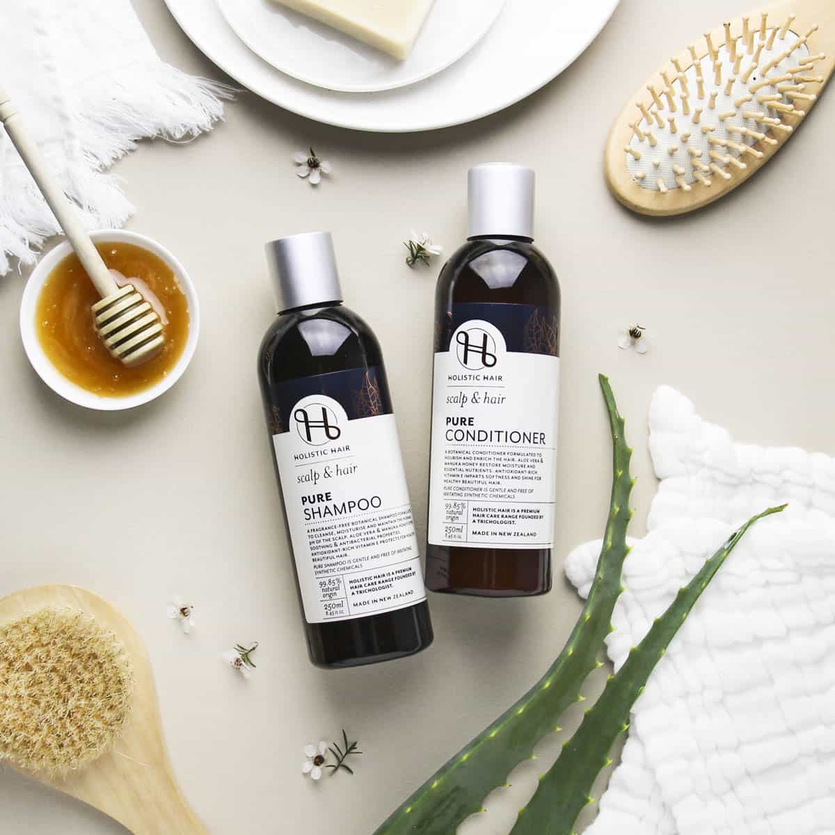 Two bottles of shampoo and conditioner with a bowl of honey, brush, and towel on a light surface.