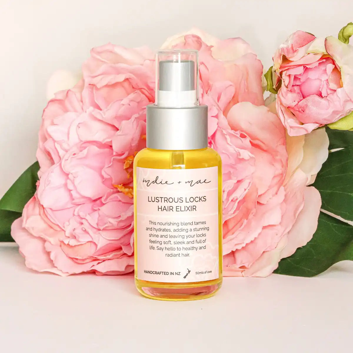 A bottle of Lustrous Locks Hair Elixir, a golden-yellow liquid in a clear bottle with a silver pump, styled in front of large pink peony flowers on a white background.