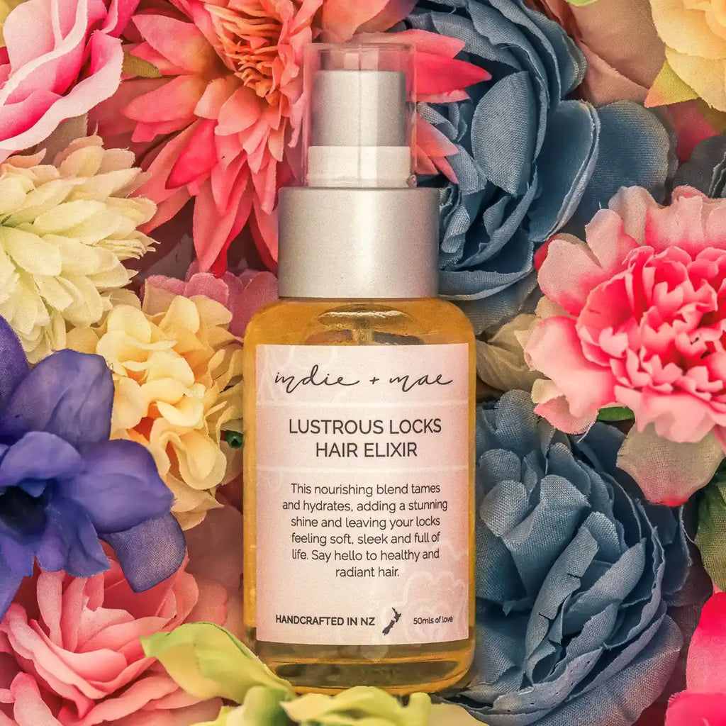 A bottle of Lustrous Locks Hair Elixir centered against a vibrant background of colorful artificial flowers, including pink, blue, yellow, and purple blossoms.