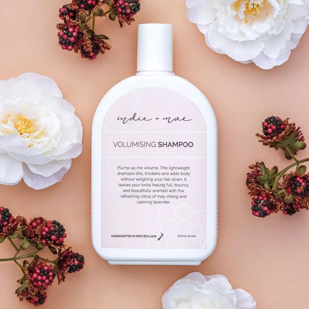 A white bottle of Indie + Moe Volumising Shampoo centered on a peach background, surrounded by white flowers and clusters of dark red berries.