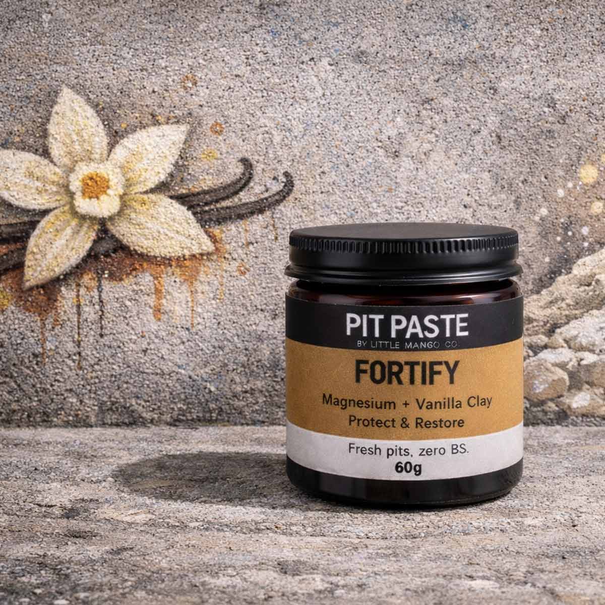 Jar of Pit Paste Fortify on a textured surface with vanilla bean and flower, clay illustrations in the background.