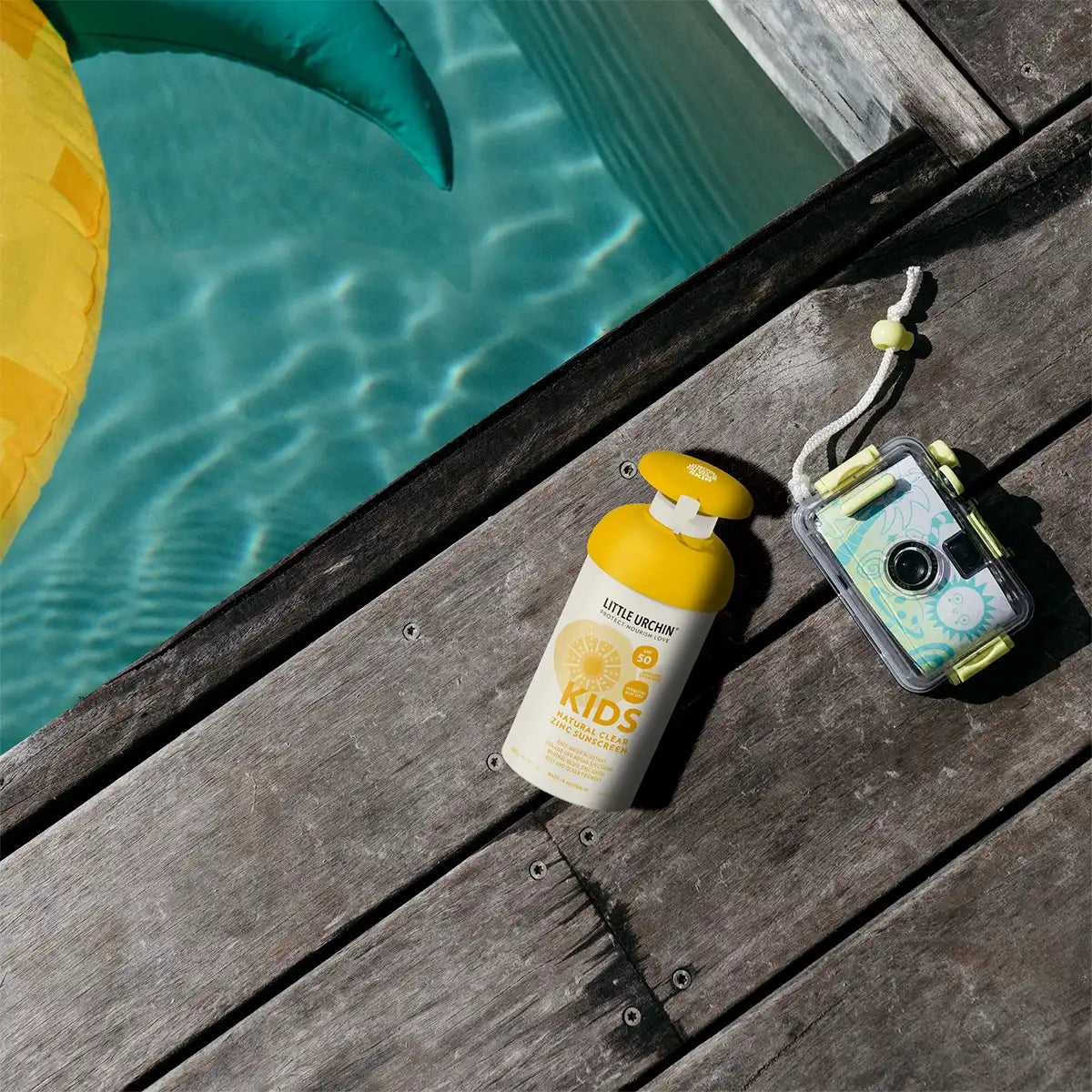A bottle of sunscreen and a waterproof camera resting on a wooden deck next to a swimming pool.