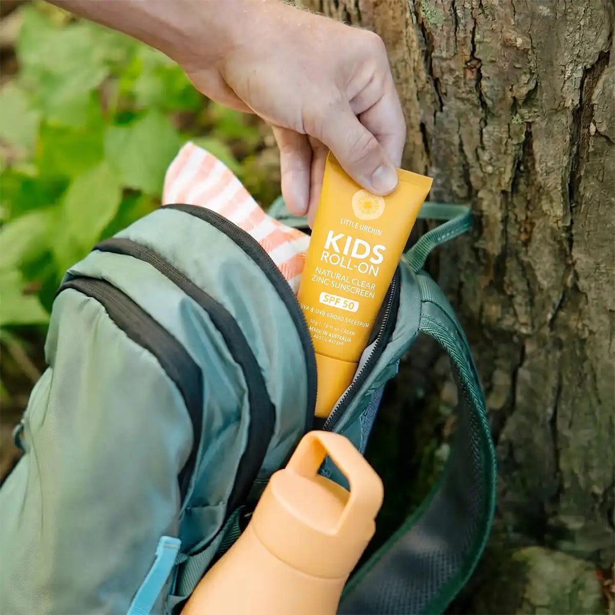 A hand places a tube of Little Urchin Roll-On Sunscreen into the pocket of a green backpack, with a water bottle and a tree trunk in the background.