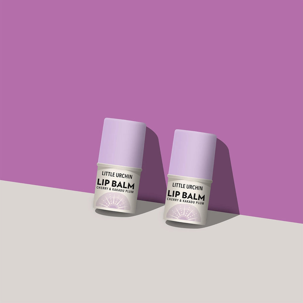 Two small lip balm tubes standing upright on a light gray surface against a purple background. The tubes are pale lavender with white labels.