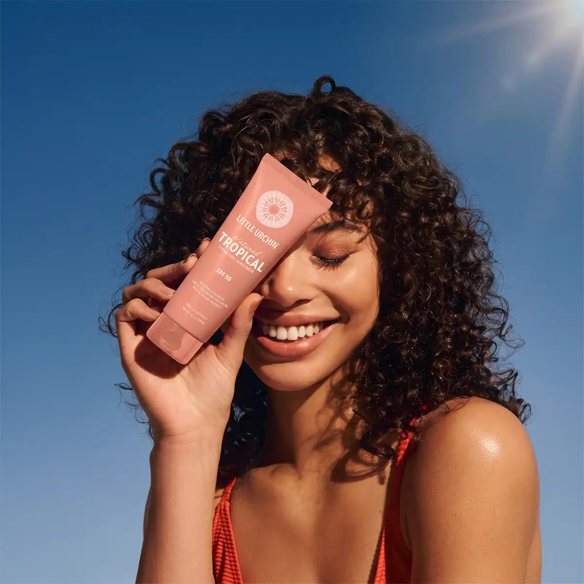 A smiling woman with curly hair holds a pink tube of Little Urchin Tropical SPF 50 near her face against a bright blue sky.
