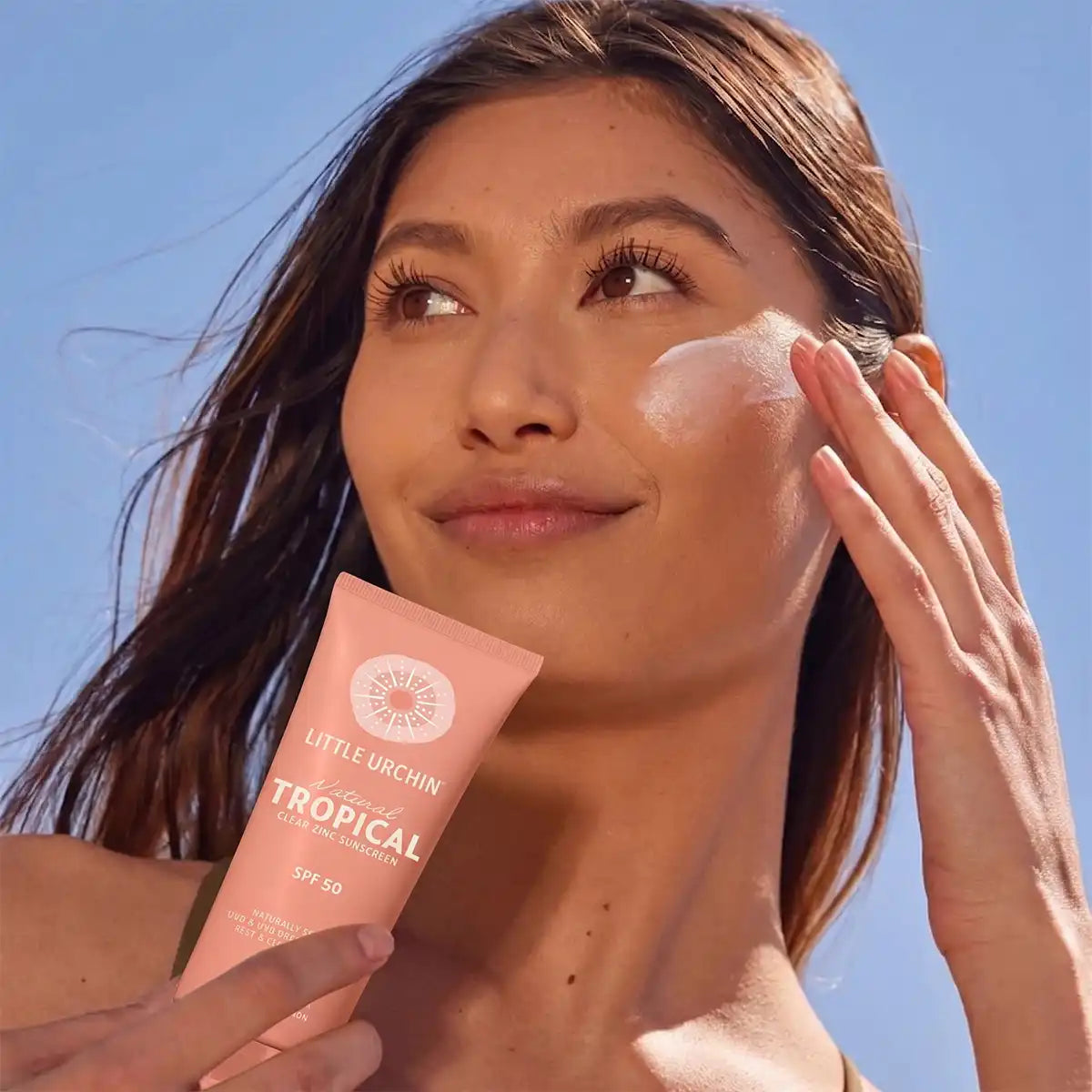 A woman outdoors under a blue sky applies white sunscreen cream to her face while holding a pink tube of Little Urchin Tropical SPF 50 sunscreen.