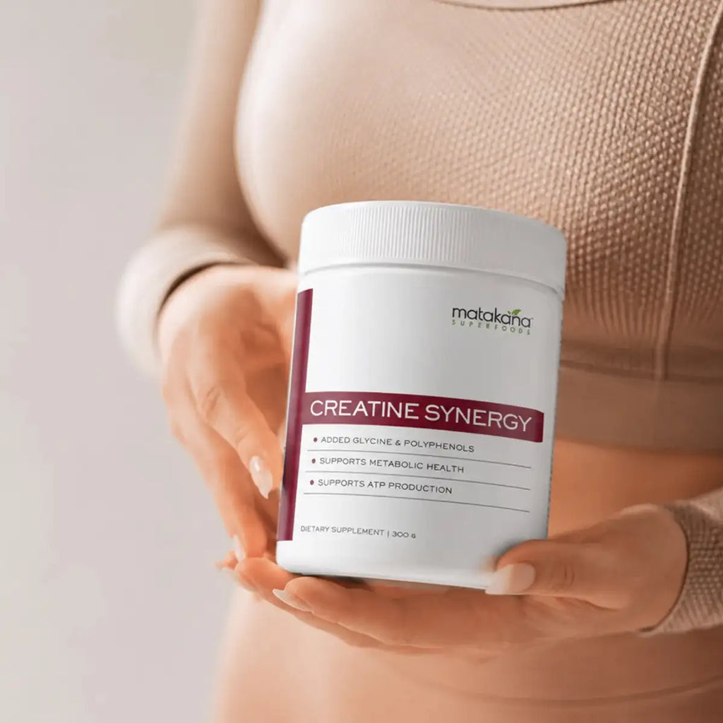 A person in a beige ribbed sweater holds a white tub of Creatine Synergy dietary supplement in both hands against a soft light background.