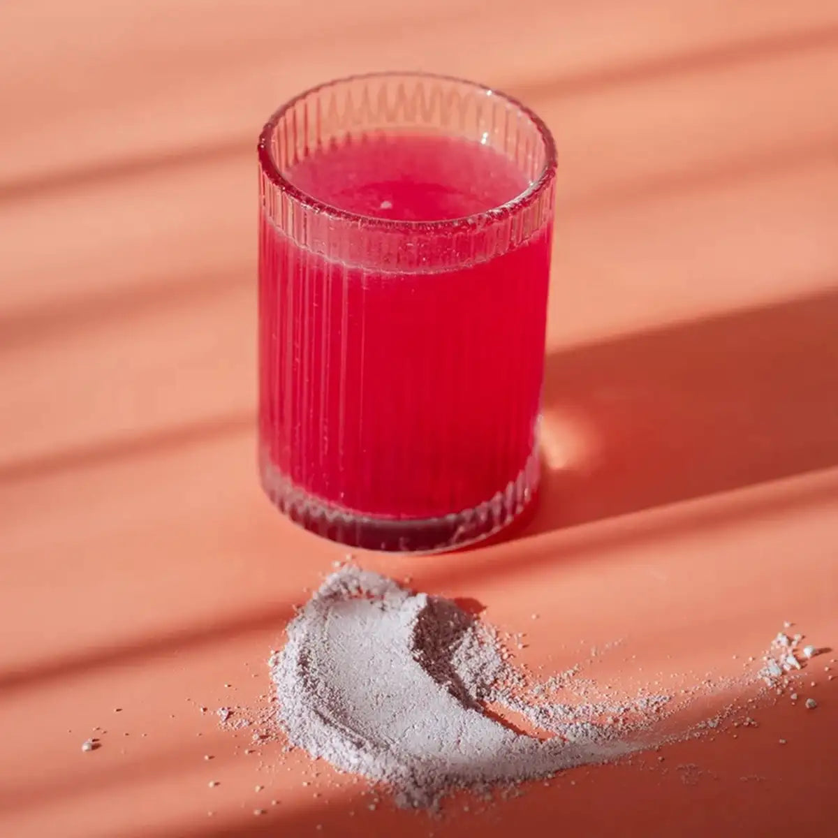 A ribbed clear glass tumbler filled with bright pink liquid, placed on a peach surface with a small pile of light gray powder in front. Strong diagonal shadows cross the scene.