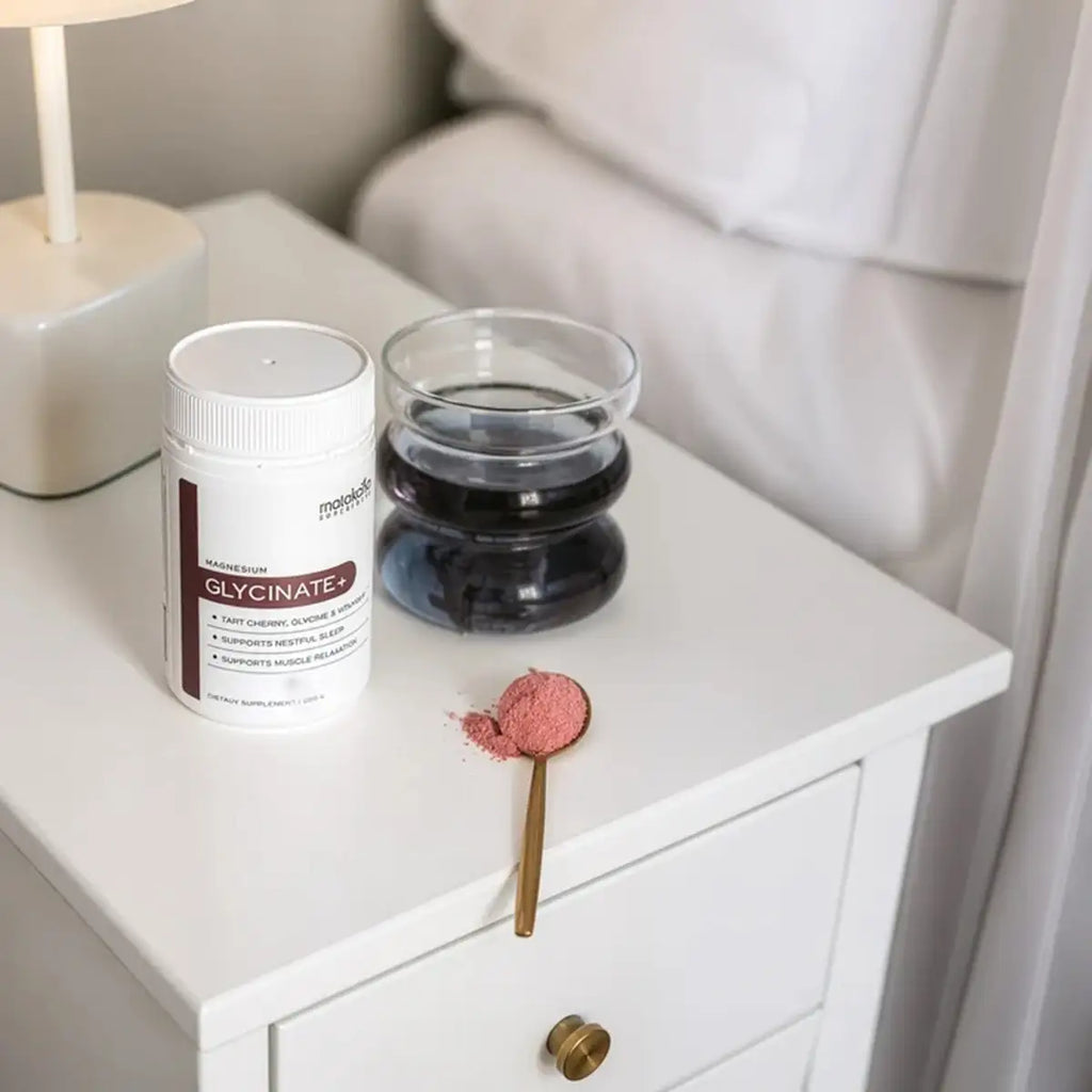 A white supplement bottle and a glass of dark powder sit on a white bedside table, with a small spoonful of pink powder nearby in a softly lit bedroom setting.