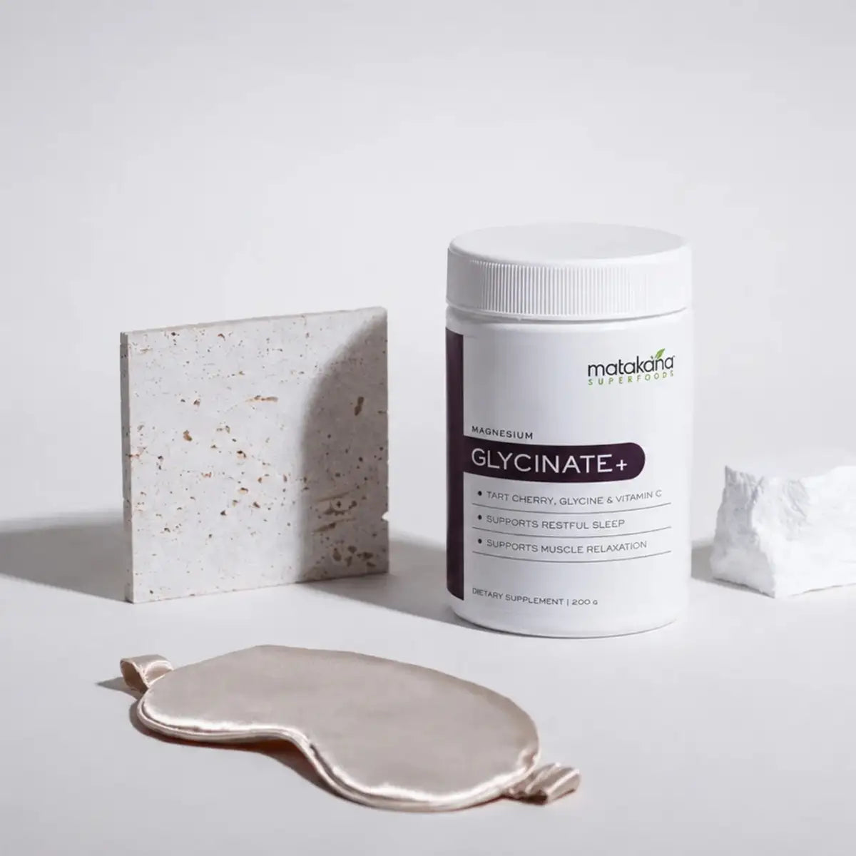 A white plastic bottle of Matakafa Supplements Glycinate+ dietary supplement stands on a light gray surface next to a square stone tile and a white rock, with a satin sleep mask in the foreground.