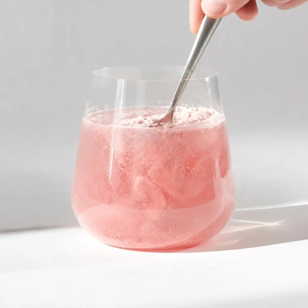 A glass filled with a pink drink and a metal spoon, held by a hand against a bright white background.