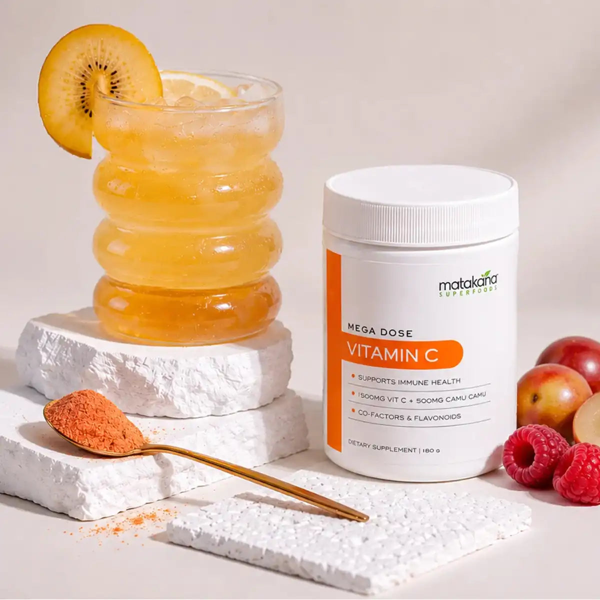 A white tub of Mega Dose Vitamin C dietary supplement stands next to a glass of golden-yellow drink garnished with a kiwi slice, with a spoon of orange powder and fruit pieces on a light beige background.