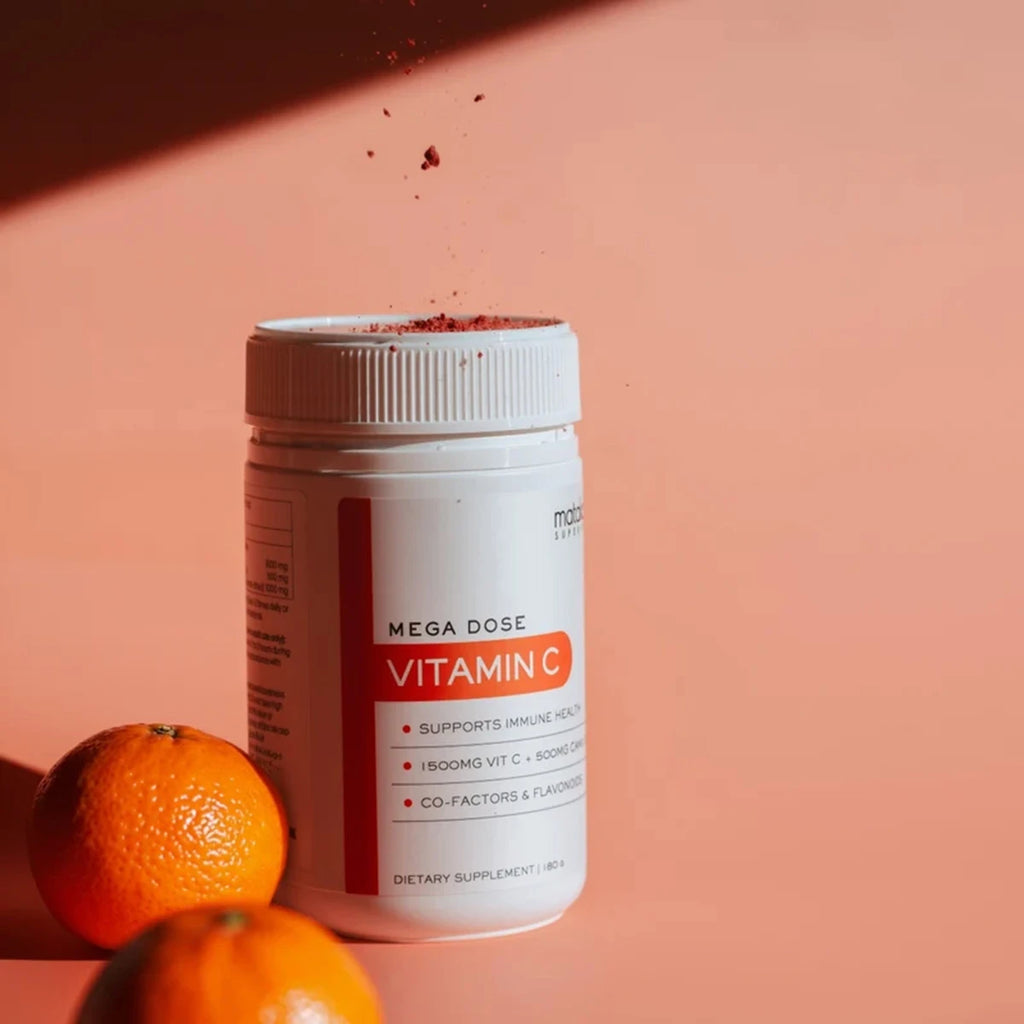 A white bottle of Mega Dose Vitamin C dietary supplement with orange accents on the label, set against a warm peach background with oranges and a sprinkle of powder in the air.