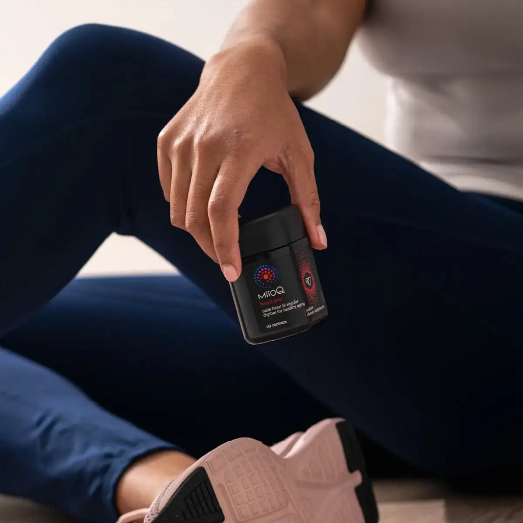 A person in dark blue leggings and a white top holds a black jar of MitoQ supplement.