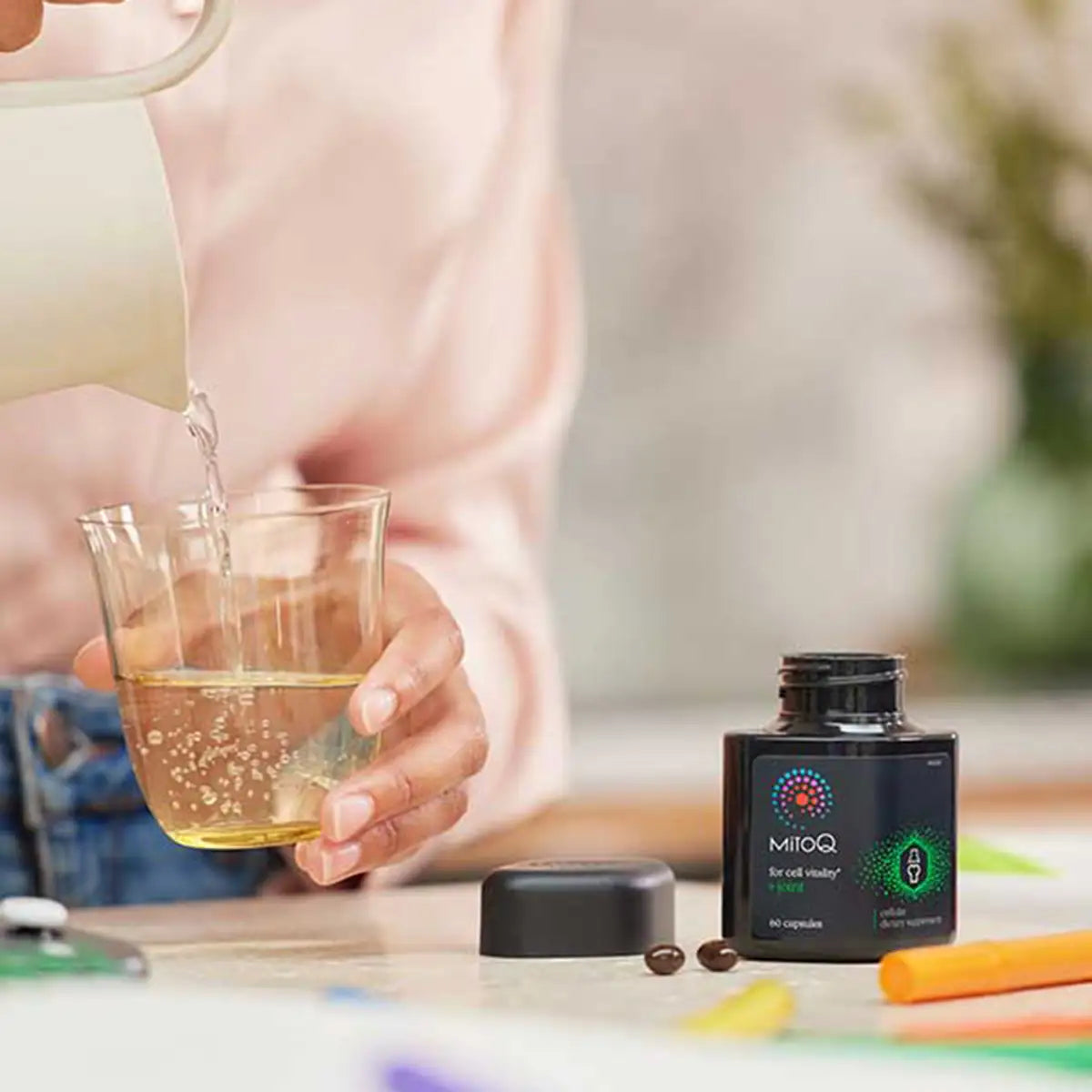 A person pouring hot water from a white pitcher into a glass beside a black supplement bottle.