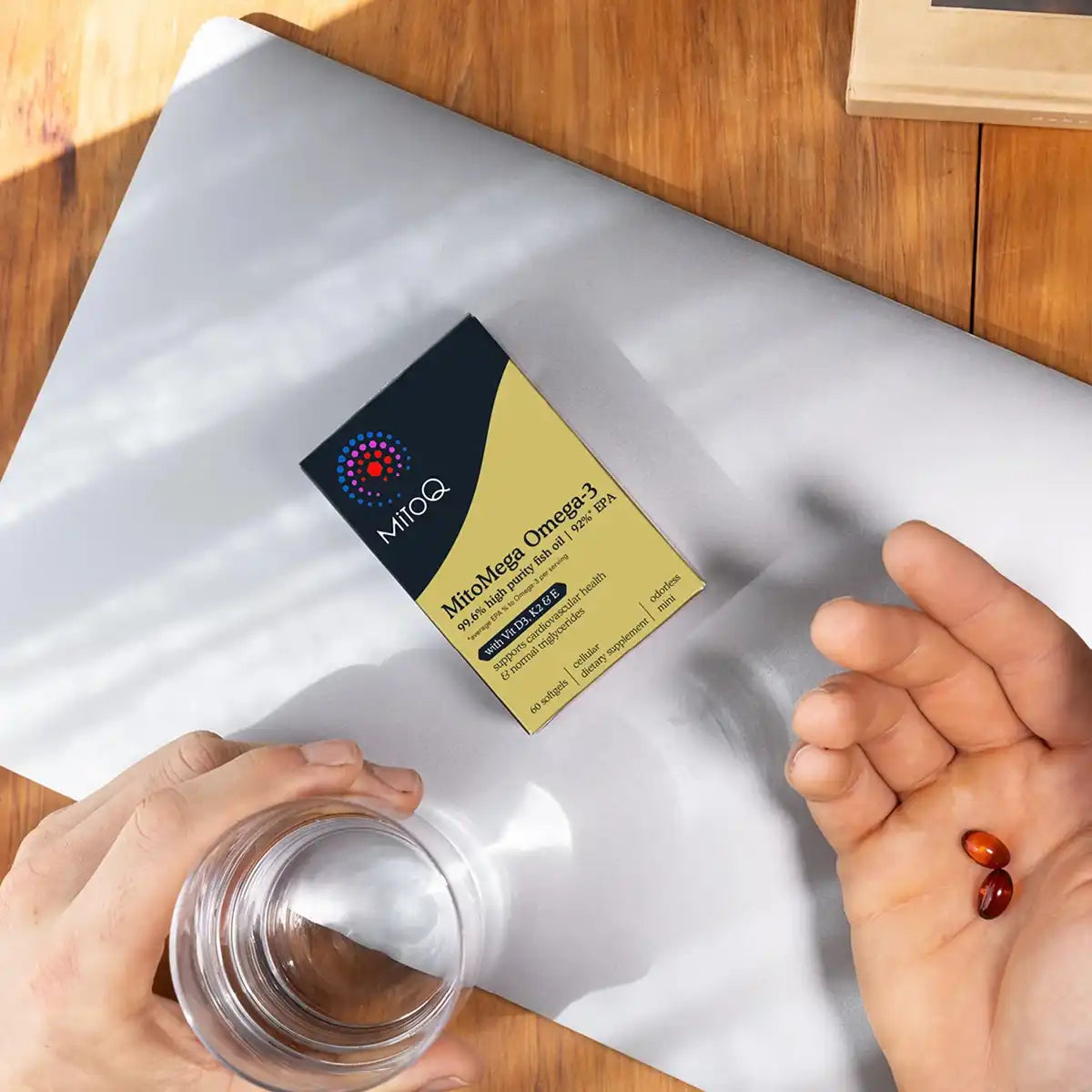 A person holds a clear glass and an open palm showing two orange-red capsules, with a box of MitoQ MitoMega Omega-3 supplement.