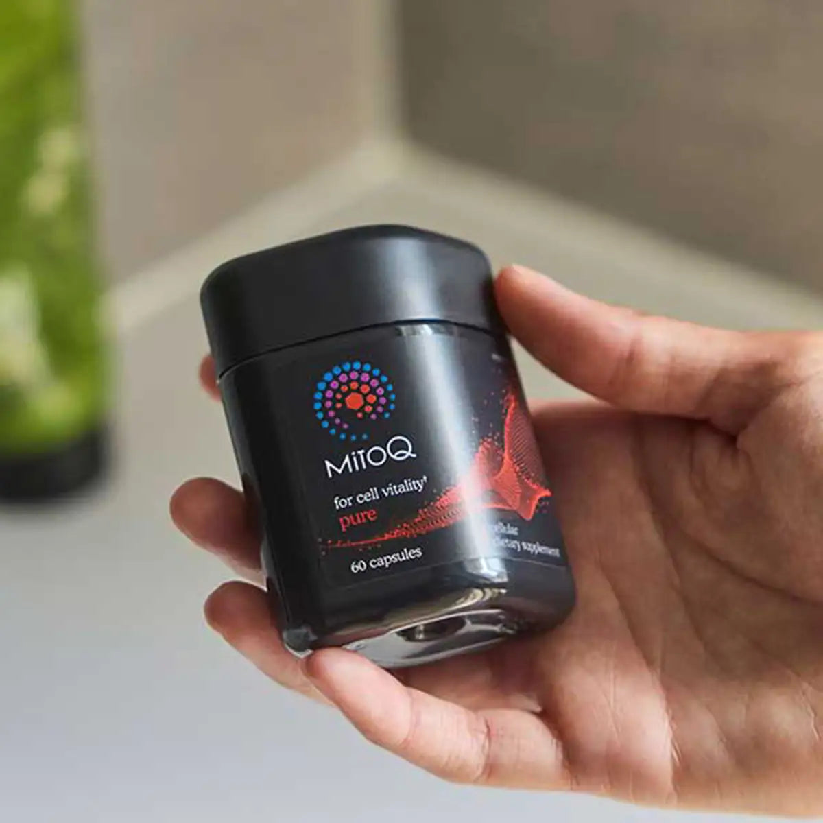 A hand holding a black jar of MitoQ dietary supplement capsules, with the brand logo and text clearly visible on the front label.