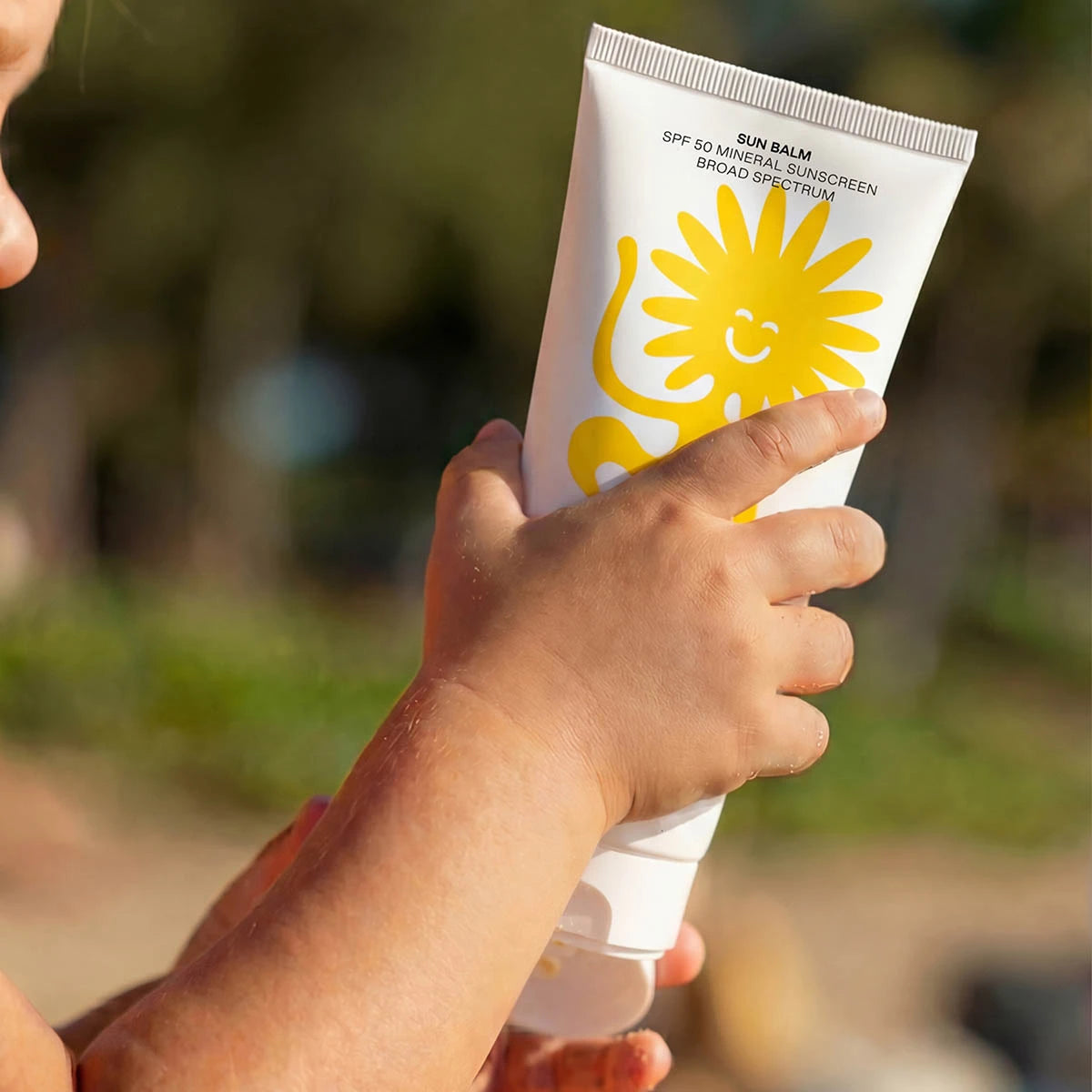 A close-up of a child holding a tube of Noody Sun Balm in an outdoor setting.