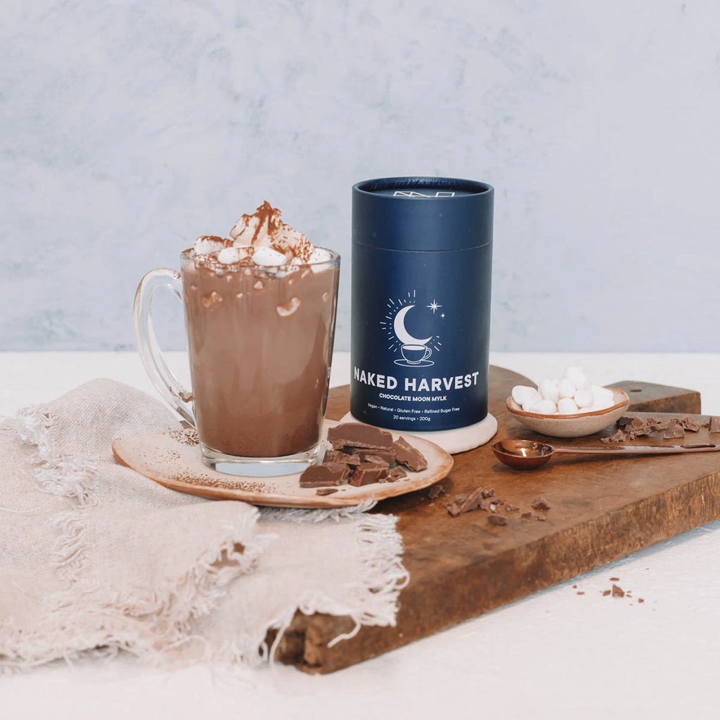 Hot chocolate with whipped cream and chocolate shavings next to a 'Naked Harvest' container on a wooden board.