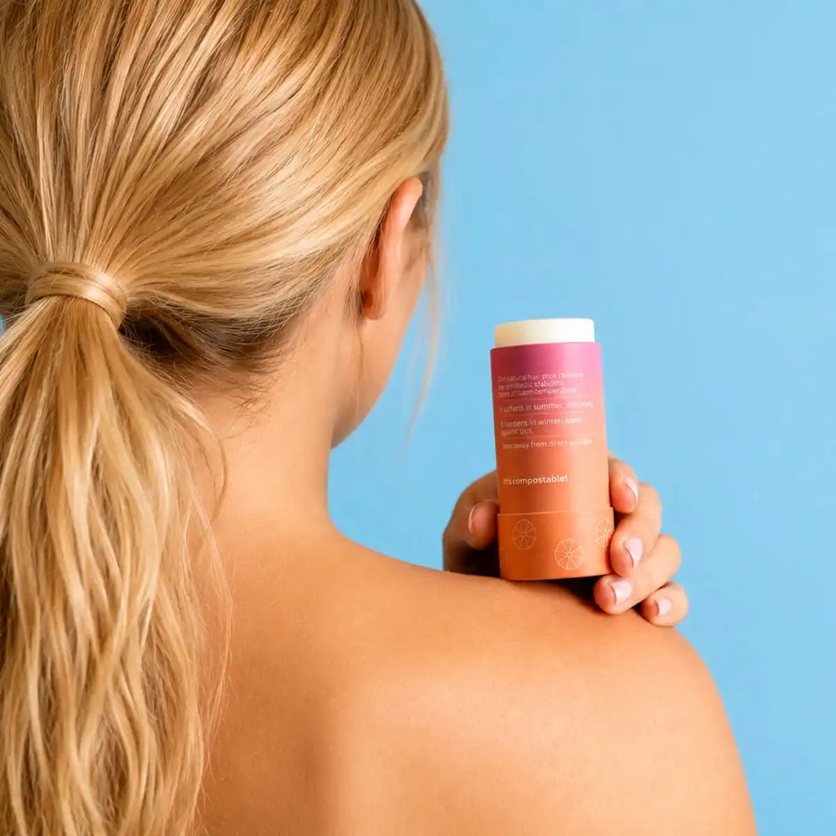 A woman with a blonde ponytail, seen from behind, holds a peach-coloured slo hair wax stick on her shoulder against a blue background