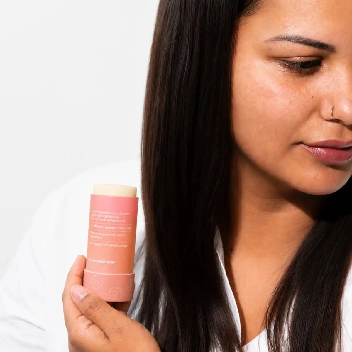 A woman with long dark hair in a white top looks down at a peach-pink slo hair wax stick held in her hand, in soft natural light