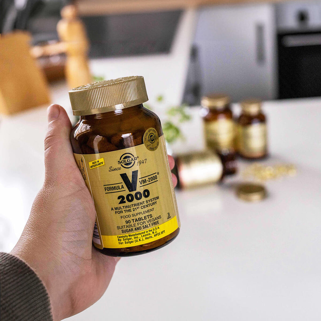 Hand holding a bottle of Solgar Formula V 2000 supplement on a kitchen counter.